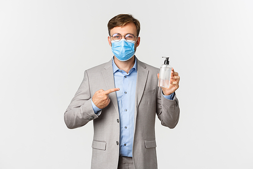 Concept of work, covid-19 and social distancing. Image of male boss in gray suit and medical mask, pointing at hand sanitizer or antiseptic, standing over white background.