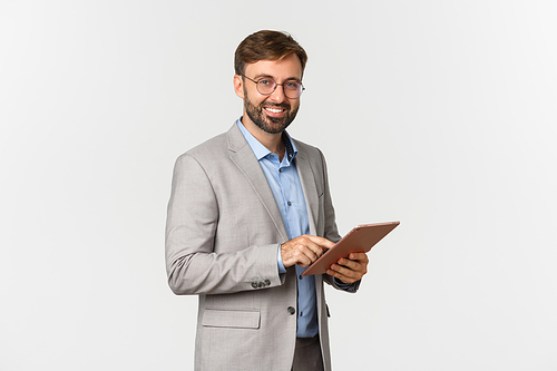 Portrait of handsome bearded male entrepreneur in grey suit and glasses, using digital tablet and working, smiling happy at camera, standing over white background.