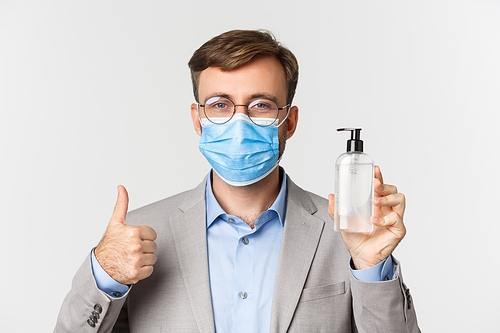 Concept of work, covid-19 and social distancing. Close-up of businessman in gray suit and medical mask, showing thumbs-up and hand sanitizer or antiseptic, standing over white background.