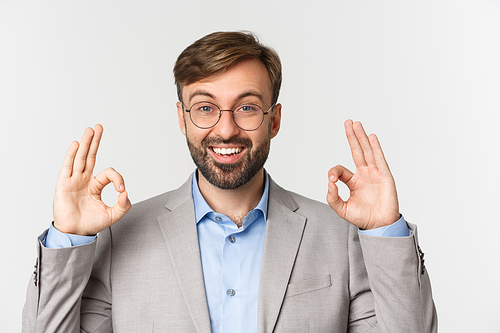 Close-up of handsome bearded man in gray suit and glasses, recommend something, showing okay sign and smiling, approve good choice, standing over white background.