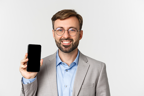 Close-up of handsome businessman in glasses and gray suit showing mobile phone screen, demonstrating an app, standing over white background.