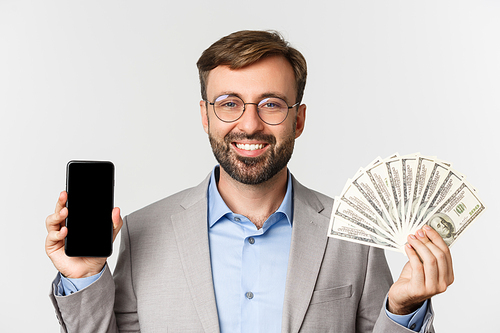 Close-up of successful businessman in gray suit and glasses, showing money and smartphone screen, standing over white background.