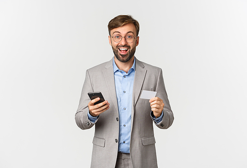 Portrait of bearded male entrepreneur in suit and glasses, looking excited while shopping online with mobile phone and credit card, standing over white background.