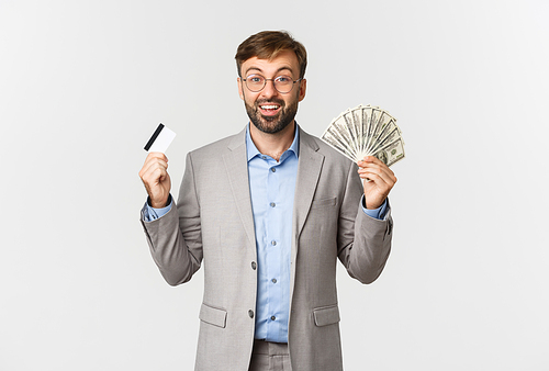 Image of handsome rich businessman in grey suit and glasses, shopping with credit card and money, smiling amazed, standing over white background.