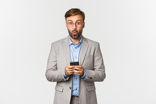 Portrait of handsome businessman with beard, wearing grey suit and glasses, looking amused after reading something interesting on mobile phone, standing over white background.