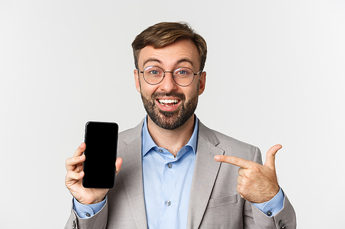 Close-up of confident businessman in glasses and gray suit, pointing finger at mobile phone screen and smiling excited, standing over white background.