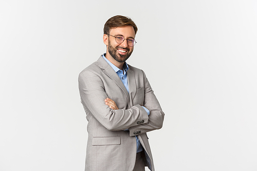 Portrait of confident businessman with beard, wearing grey suit and glasses, cross arms on chest and smiling self-assured, standing over white background.