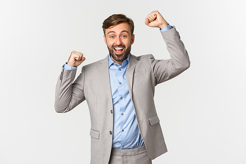 Portrait of successful smiling businessman with beard, wearing grey suit, cheering and celebrating victory, saying yes or hooray, triumphing and winning, standing over white background.