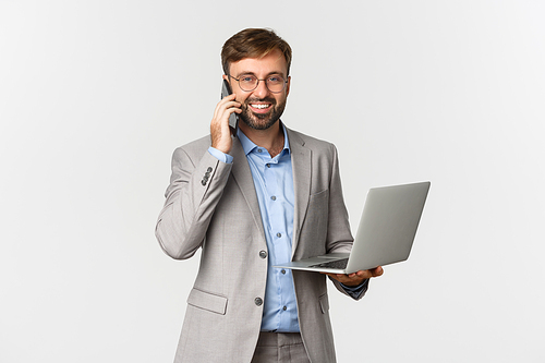 Image of successful and confident businessman in glasses and grey suit, talking on phone and holding laptop, smiling happy at camera, standing over white background.