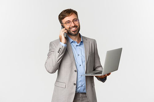 Image of successful and confident businessman in glasses and grey suit, talking on phone and holding laptop, smiling pleased at camera, standing over white background.