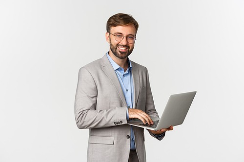 Portrait of successful and confident businessman in grey suit and glasses, working with laptop and smiling happy at camera, standing over white background.