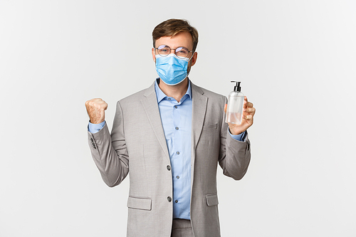 Concept of work, covid-19 and social distancing. Image of cheerful and confident businessman encourage employees to use hand sanitizer or antiseptic, standing in medical mask and gray suit.