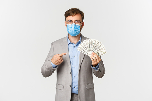 Concept of business, covid-19 and social distancing. Portrait of successful businessman in medical mask earn money, pointing finger at cash, standing over white background.