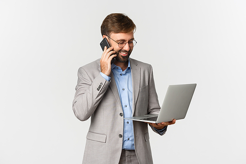 Image of successful businessman talking on phone and looking satisfied at laptop screen, working over white background.