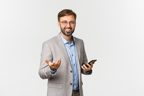 Portrait of confident businessman with beard, wearing glasses and grey suit, holding mobile phone and pointing at you pleased, standing over white background.