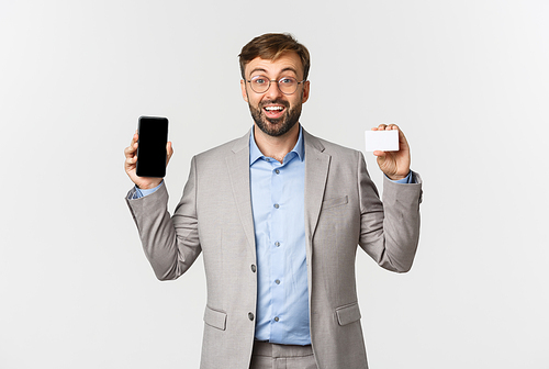 Cheerful male entrepreneur with beard, wearing grey suit and glasses, showing app on mobile phone screen and credit card, smiling excited, standing over white background.