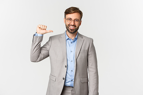 Portrait of confident and successful businessman with beard, wearing grey suit and glasses, pointing at himself and smiling, standing over white background.