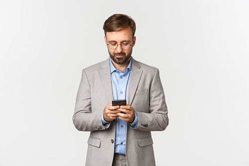 Handsome caucasian businessman in glasses and gray suit, writing message on smartphone, looking at mobile phone, standing over white background.