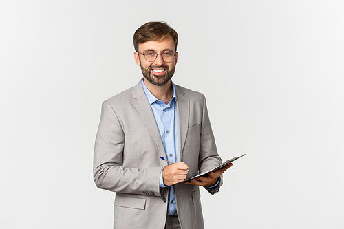 Portrait of successful and confident businessman in gray suit and glasses, writing something on clipboard and smiling, inspecting work environment.