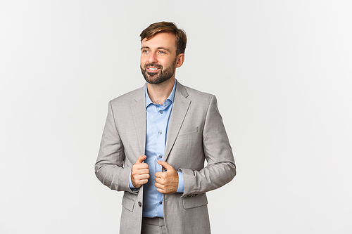 Image of successful and confident businessman in gray suit and blue shirt, smiling pleased and looking left, standing over white background.