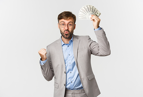 Successful bearded businessman in grey suit and glasses, winning cash, holding money and triumphing, standing happy over white background.