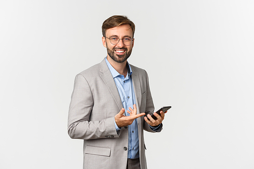 Portrait of handsome bearded businessman in grey suit and glasses, holding mobile phone, pointing at smartphone and smiling, standing over white background.