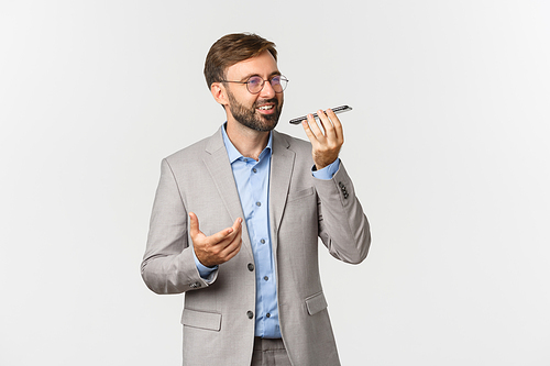 Handsome smiling businessman with glasses and gray suit, talking on speakerphone, holding mobile phone near mouth and smiling, recording voice message, white background.