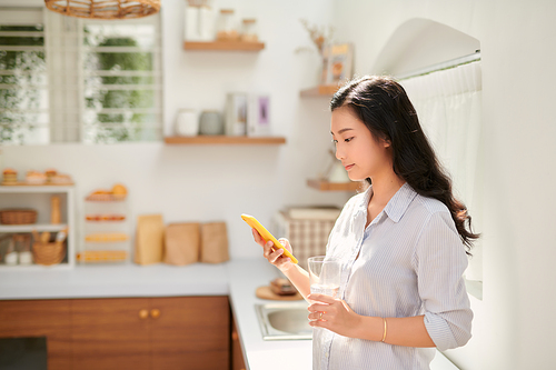 Beautiful happy girl drinking water while using mobile phone at home kitchen