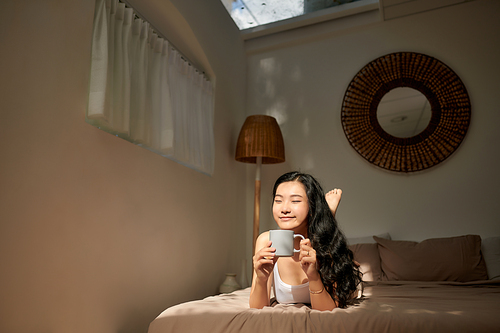 Young woman in beige pajamas lying on the bed at home.Relaxing and drinking cup of hot coffee or tea.