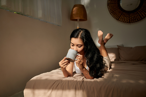 beautiful young woman hold cup of coffee lying think relaxing on the bed, happy smile day dreaming with tea mug in hands
