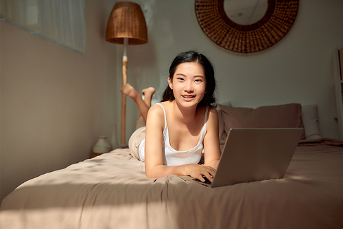 Happy casual beautiful woman working on a laptop lying on the bed in the house.