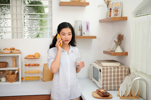 Portrait of smiling young beautiful woman standing, drinking water and talking on smartphone in kitchen.