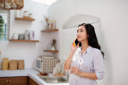 Portrait of smiling young beautiful woman standing, drinking water and talking on smartphone in kitchen.
