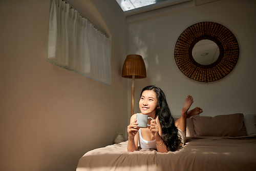 beautiful young woman hold cup of coffee lying think relaxing on the bed, happy smile day dreaming with tea mug in hands