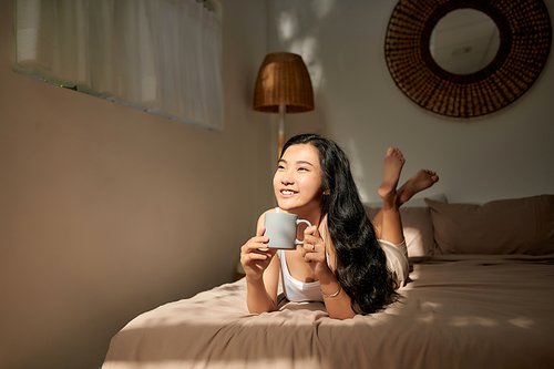 Young woman in beige pajamas lying on the bed at home.Relaxing and drinking cup of hot coffee or tea.