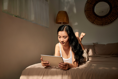 A smiling woman lies on the bed scrolling through her tablet.