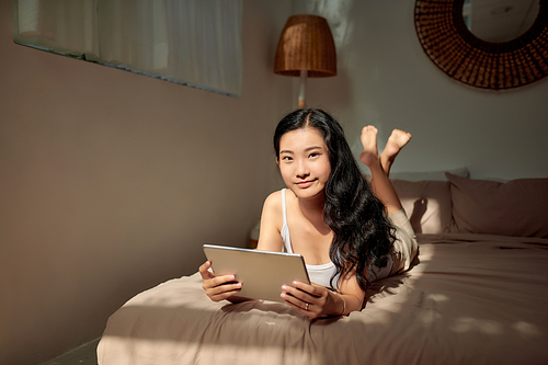 A smiling woman lies on the bed scrolling through her tablet.