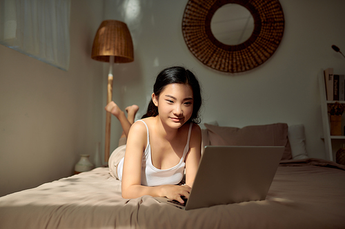 Beautiful young woman relaxing on her bed working with her laptop