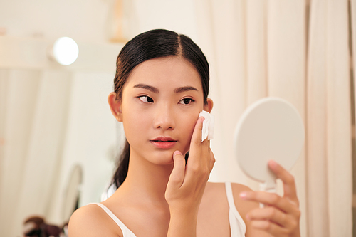 Final touches. Side view of beautiful young woman using cotton disk and looking at her reflection in mirror while sitting at the dressing table