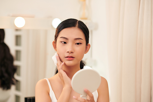 Young cute woman remove makeup , cleaning face with cotton pads looking in mirror