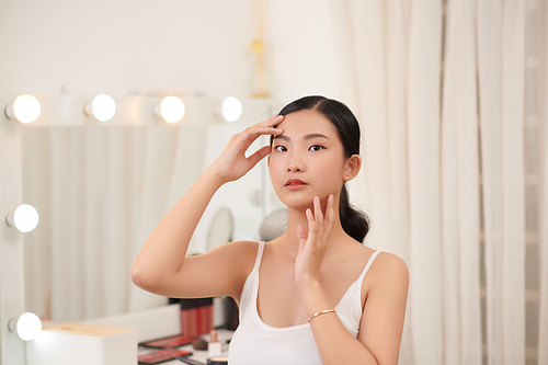 Beautiful young woman is touching her face and smiling while looking at the mirror