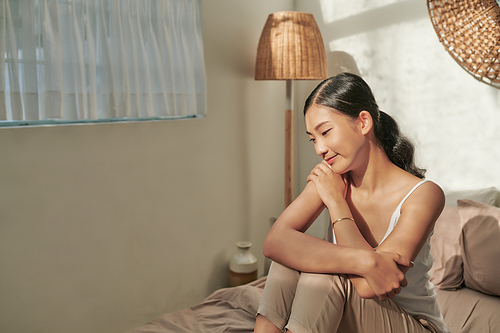 Cheerful happy asian girl in sleepwear smiling, sitting on bed at home.