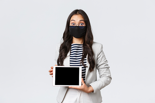 Business, finance and employment, covid-19 preventing virus and social distancing concept. Excited asian businesswoman in face mask and glasses widen eyes surprised, showing digital tablet display.