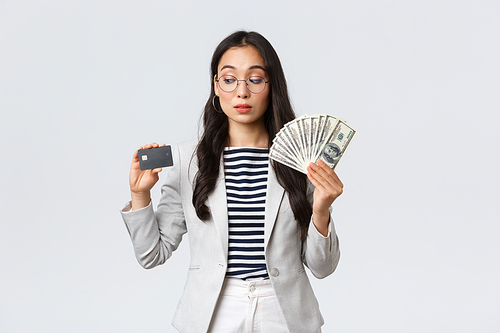 Business, finance and employment, entrepreneur and money concept. Indecisive cute asian office lady thinking about putting cash on deposit, looking curious at credit card, white background.
