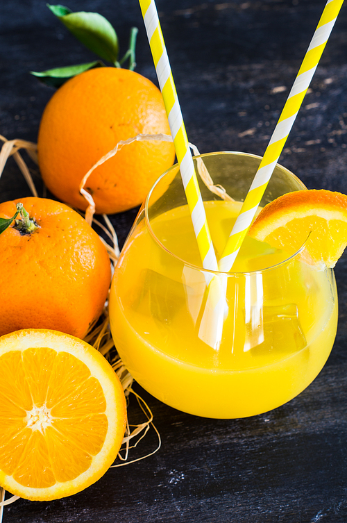 Glass of orange juice with straw and ripe orange fruits with green leaves on dark rustic wooden table