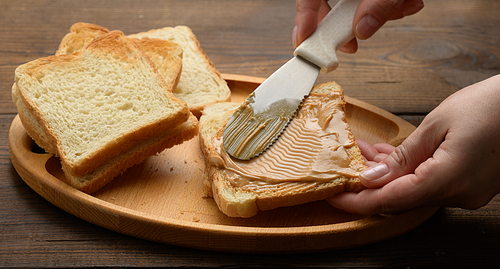 female hand spreads peanut butter on a square slice of white wheat flour, breakfast