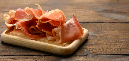 sliced thin slices of prosciutto on wooden brown board, wooden table background