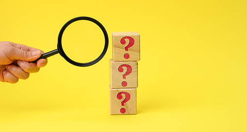 wooden cubes with question marks and a female hand holds a magnifying glass on a yellow background. The concept of finding answers to unknown questions, solving a problem. Find information