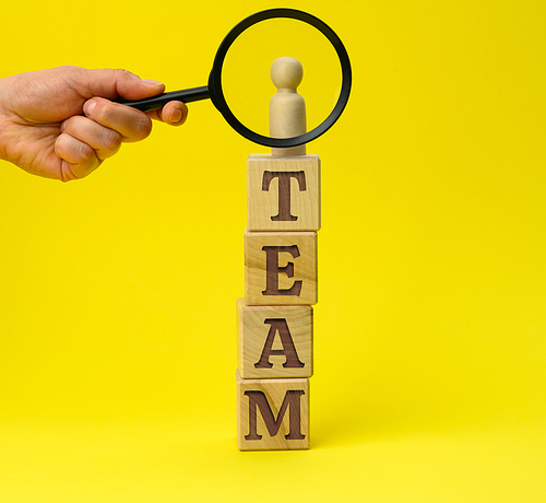 wooden figurine of a man on a pedestal and a hand with a magnifying glass on a yellow background. The concept of recruiting and finding talented employees, close supervision. Team building