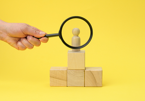 wooden figurine of a man on a pedestal and a hand with a magnifying glass on a yellow background. The concept of recruiting and finding talented employees, close supervision. Team building